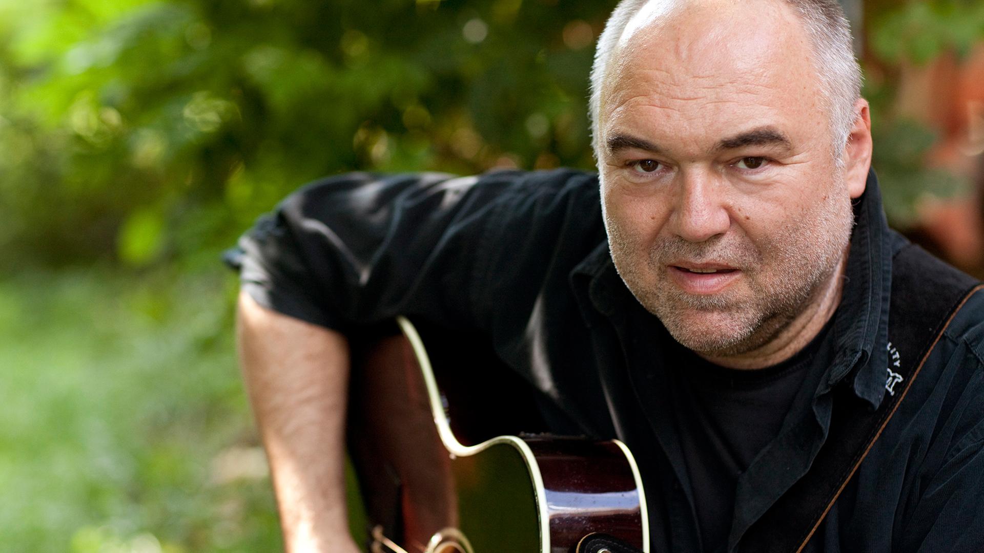 Zu sehen ist der oberfränkische Songpoet Wolfgang Buck sitzend und in die Kamera blickend mit einer Gitarre spielbereit unter dem rechten Arm. Zu sehen ist der oberfränkische Songpoet Wolfgang Buck sitzend und in die Kamera blickend mit einer Gitarre spielbereit unter dem rechten Arm.