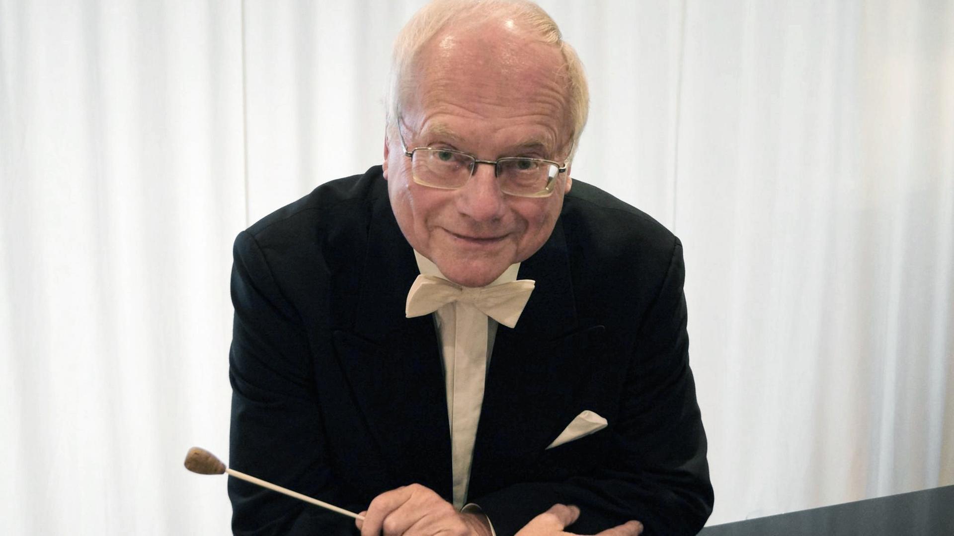 Ein älterer Mann mit Brille und weißem Haar lächelt in die Kamera, während er sich mit seinen Ellenbogen auf einen Flügel stützt und in einer Hand einen Dirigierstock hält. Ein älterer Mann mit Brille und weißem Haar lächelt in die Kamera, während er sich mit seinen Ellenbogen auf einen Flügel stützt und in einer Hand einen Dirigierstock hält.