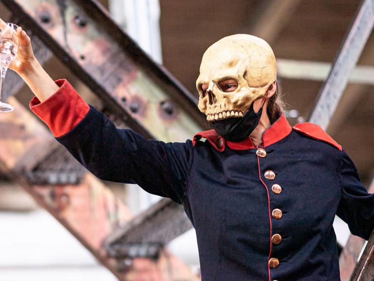 Eine Person in Uniform und mit Totenschädelmaske erhebt feierlich ein Glas