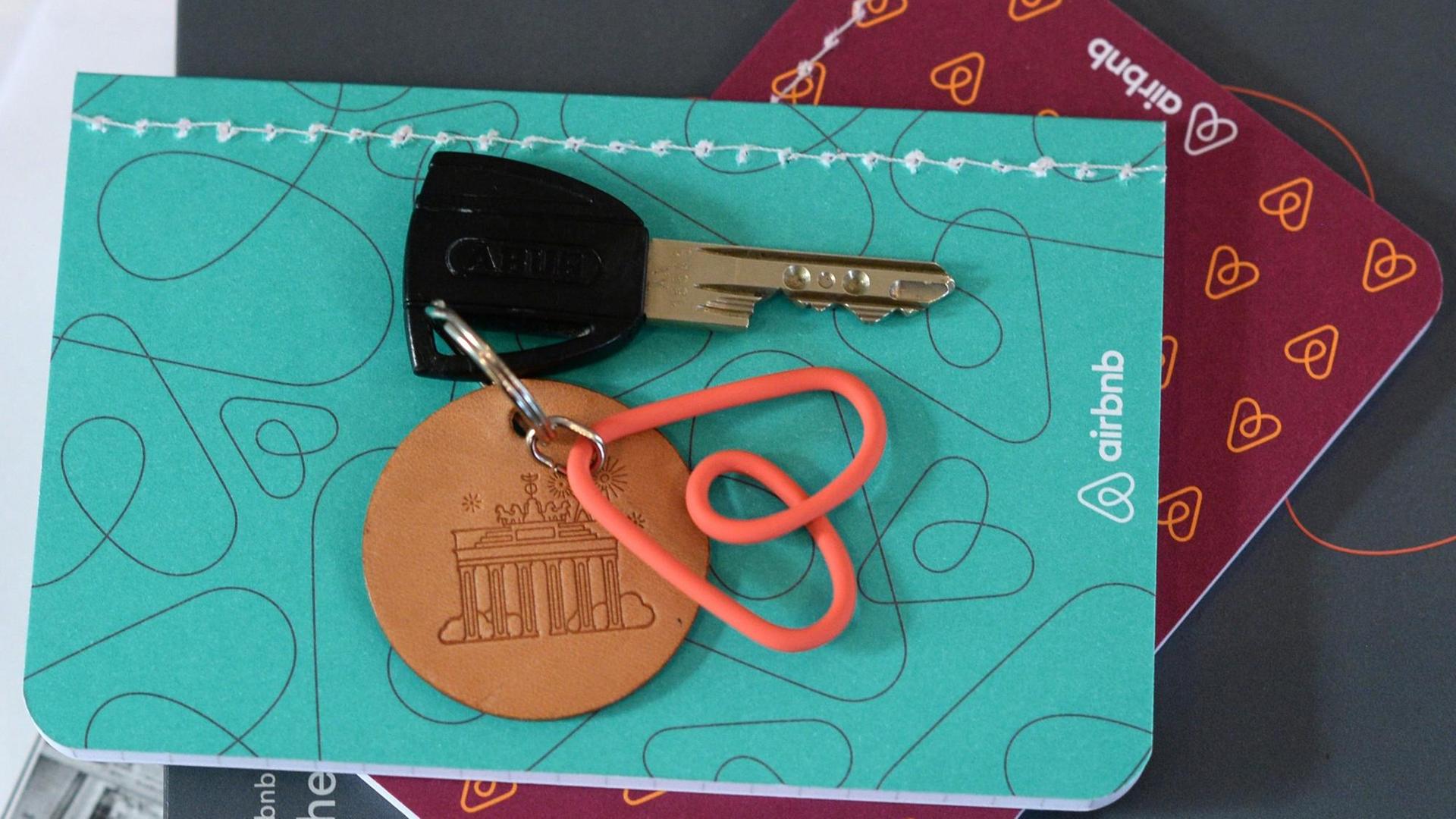 Ein Schlüsselanhänger mit Schlüssel mit dem Logo der Online-Plattform Airbnb liegt auf einem Tisch und einem Notizbuch in einer Ferienwohnung.