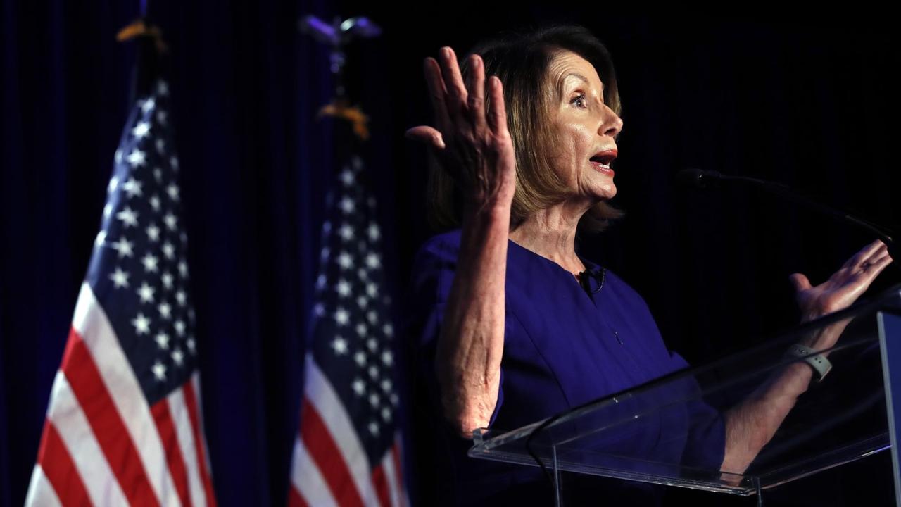 Pelosi steht an einem Rednerpult, spricht und hebt gestikulierend beide Hände. Im dunklen Hintergrund sieht man zwei US-Fahnen. Pelosi steht an einem Rednerpult, spricht und hebt gestikulierend beide Hände. Im dunklen Hintergrund sieht man zwei US-Fahnen.
