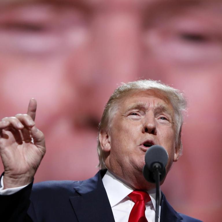 Donald Trump mit erhobenem Zeigefinger vor einem großen Bild von ihm selbst