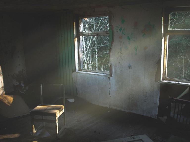 Blick in ein Haus, wo niemand mehr wohnt. Die Fenster sind kaputt, in der Mitte des Raumes steht ein Stuhl.