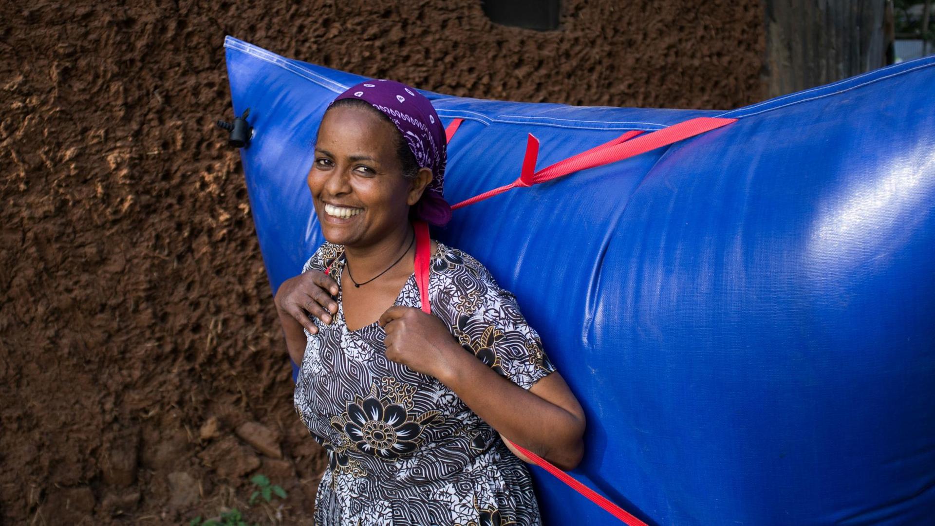 Eine Afrikanerin im geblümten Kleid trägt einen riesigen knallblauen Plastikrucksack voller Biogas auf dem Rücken.