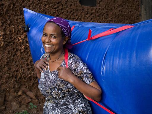 Eine Afrikanerin im geblümten Kleid trägt einen riesigen knallblauen Plastikrucksack voller Biogas auf dem Rücken.