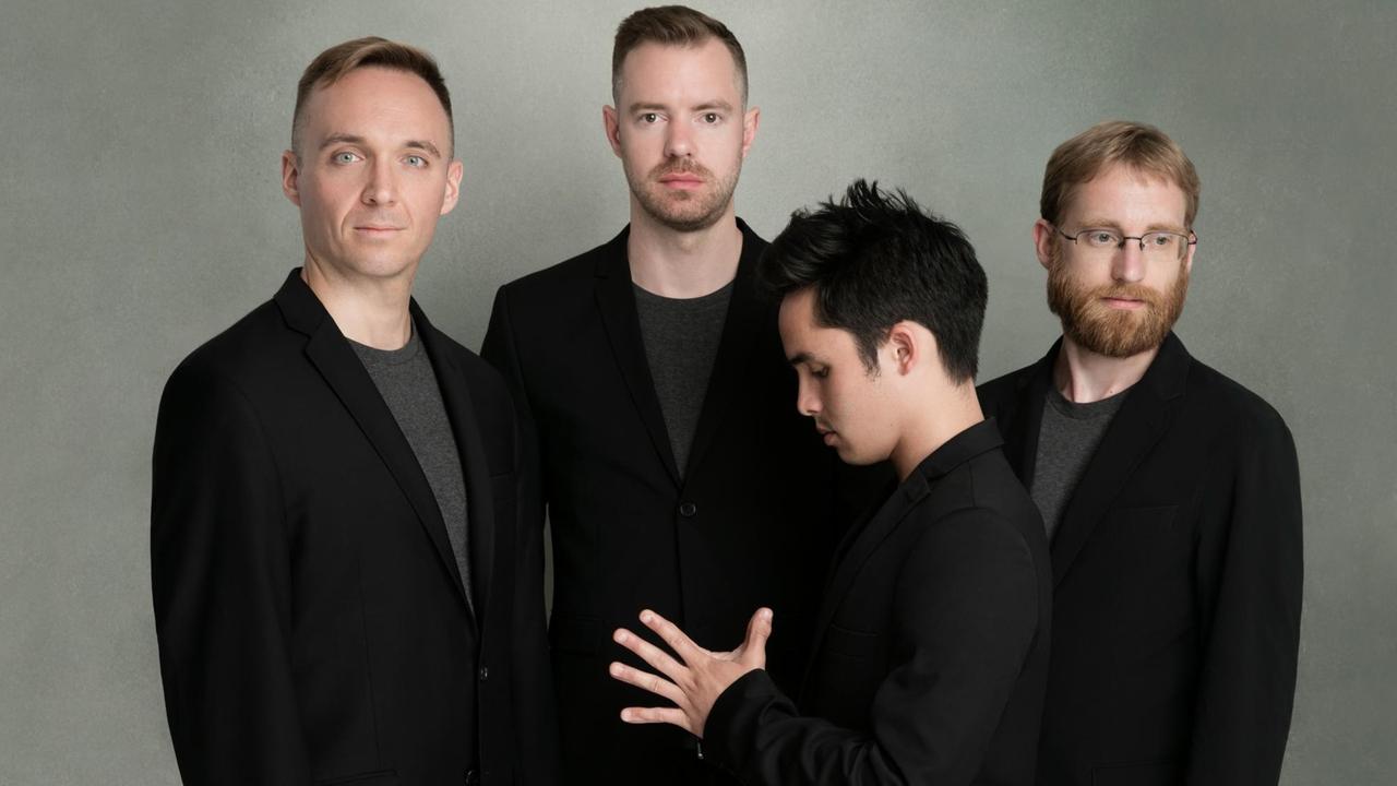 Die Vier Musiker stehen in dunkler Kleidung dicht beieinander.