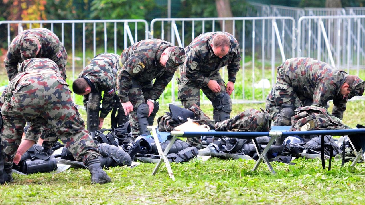 Die Soldaten in Tarnuniformen legen auf einer Wiese ihren Beinschutz an. Um sie herum liegt ihr Gepäck. Hinter ihnen sind Absperrungen zu sehen, davor ein Feldbett. Die Soldaten in Tarnuniformen legen auf einer Wiese ihren Beinschutz an. Um sie herum liegt ihr Gepäck. Hinter ihnen sind Absperrungen zu sehen, davor ein Feldbett.