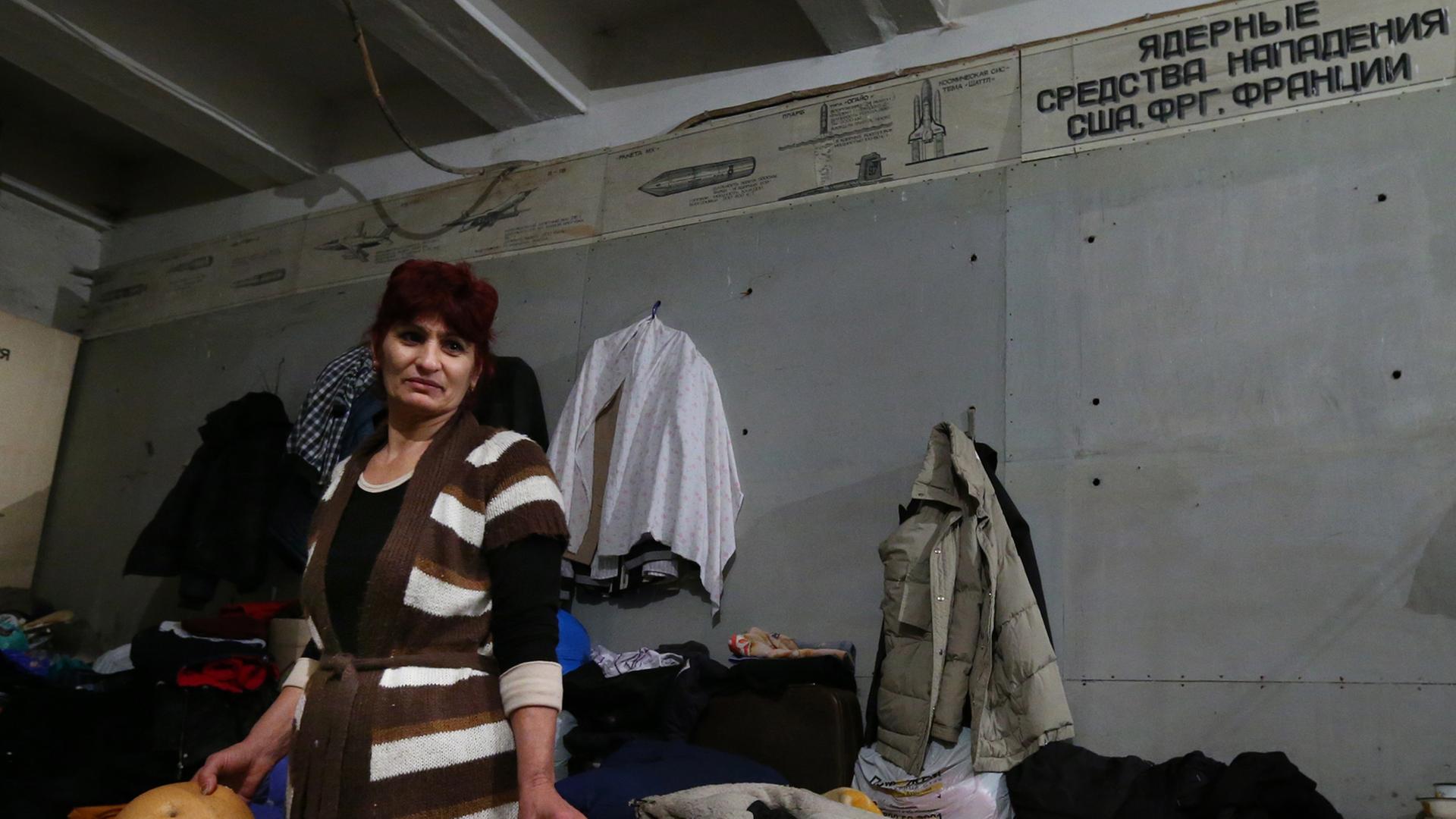 Eine Frau steht mit mehreren großen Broten in den Händen in einem Luftschutzbunker, hinter sind eine Matratze und Kleidung zu sehen Eine Frau steht mit mehreren großen Broten in den Händen in einem Luftschutzbunker, hinter sind eine Matratze und Kleidung zu sehen