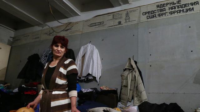 Eine Frau steht mit mehreren großen Broten in den Händen in einem Luftschutzbunker, hinter sind eine Matratze und Kleidung zu sehen