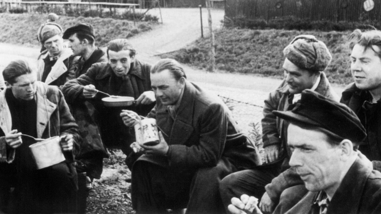 Kriegsheimkehrer nehmen im Lager Friedland eine Mahlzeit zu sich, im Hintergrund die als Nissenhütten bezeichneten Unterkünfte.