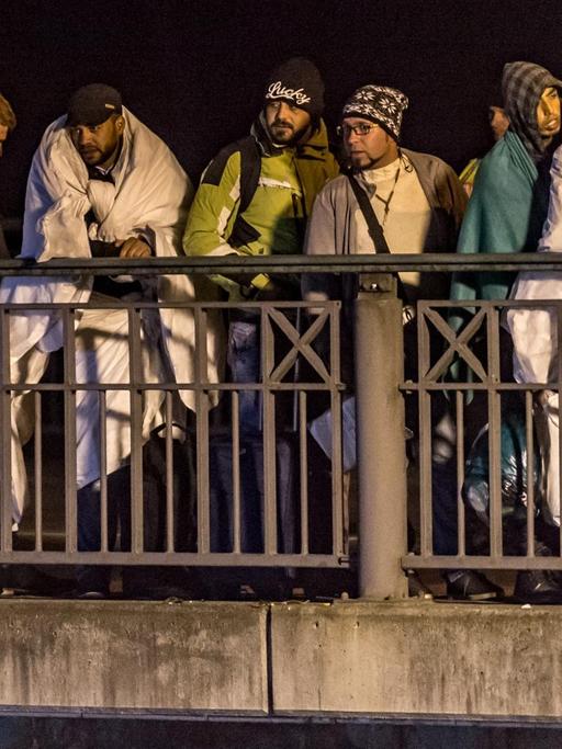 Flüchtlinge stehen nachts in Decken gehüllt auf einer Brücke.