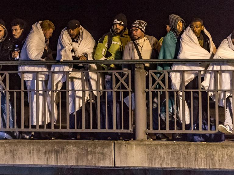 Flüchtlinge stehen nachts in Decken gehüllt auf einer Brücke.