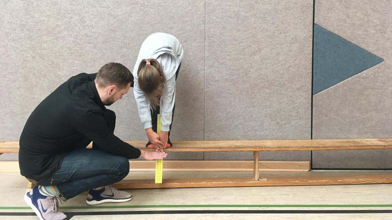 Ein Erwachsener hält ein Zollstock an eine Bank, darauf steht ein Mädchen, das den Zollstock anfässt. Ein Erwachsener hält ein Zollstock an eine Bank, darauf steht ein Mädchen, das den Zollstock anfässt.
