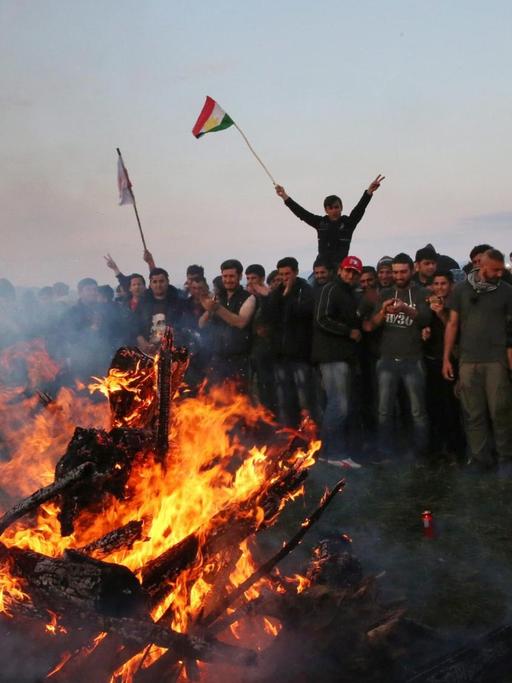 Menschen stehen um ein Feuer in Idomeni herum, es wehen kurdische Flaggen, das kurdische Neujahrsfest wird gefeiert.