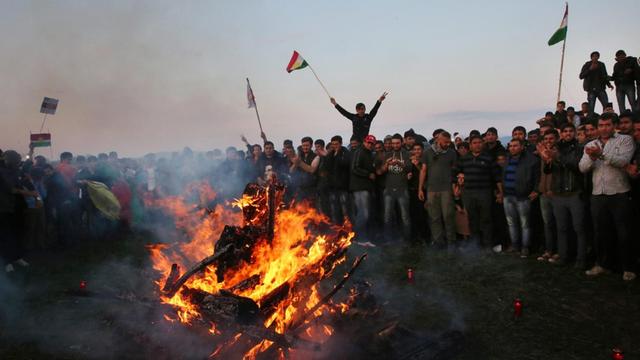 Menschen stehen um ein Feuer in Idomeni herum, es wehen kurdische Flaggen, das kurdische Neujahrsfest wird gefeiert.