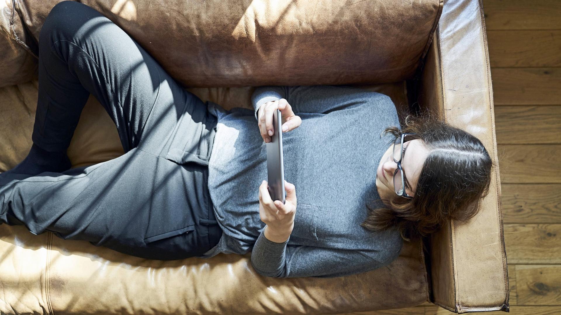 Eine Frau liegt auf dem Sofa und schaut auf den Bildschirm ihres Tablets. Eine Frau liegt auf dem Sofa und schaut auf den Bildschirm ihres Tablets.