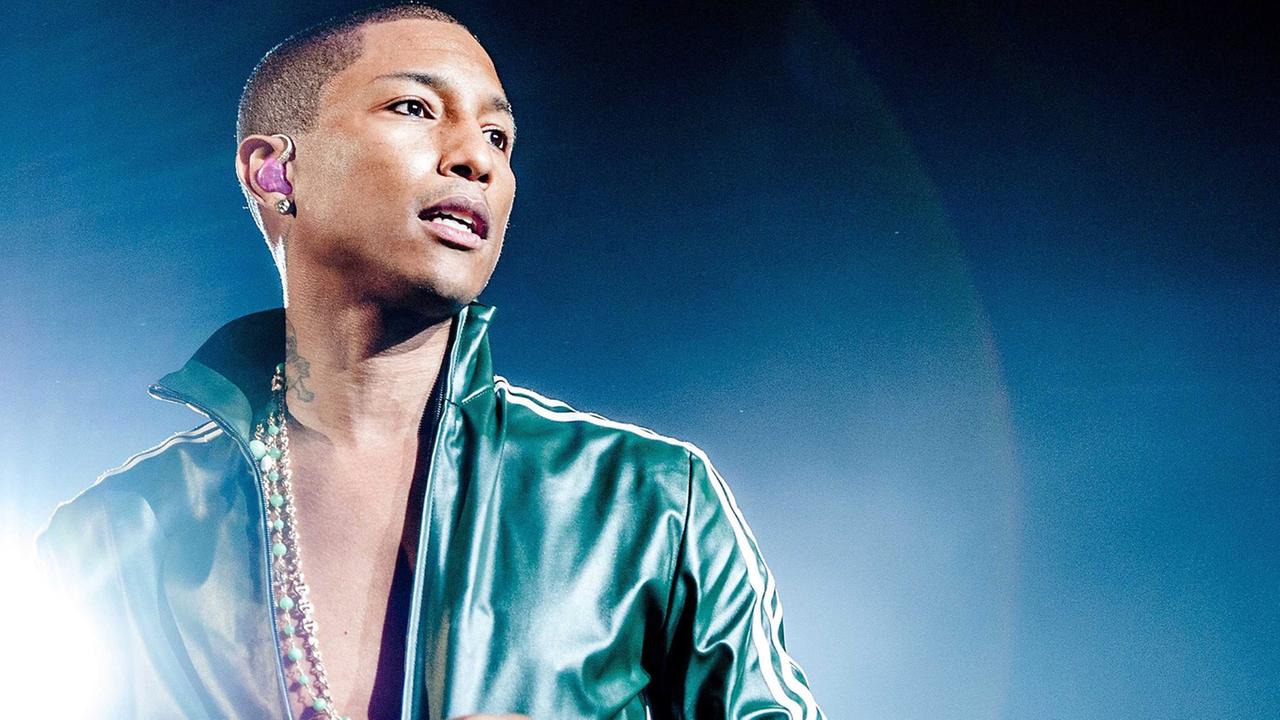 Pharrell Williams in grüner Jacke live auf einer Bühne; neben ihm strahlt ein Scheinwerfer in die Kamera. Die Jacke ist fast bis zum Bauchnabel offen. Willimas trägt eine lange Goldkette.