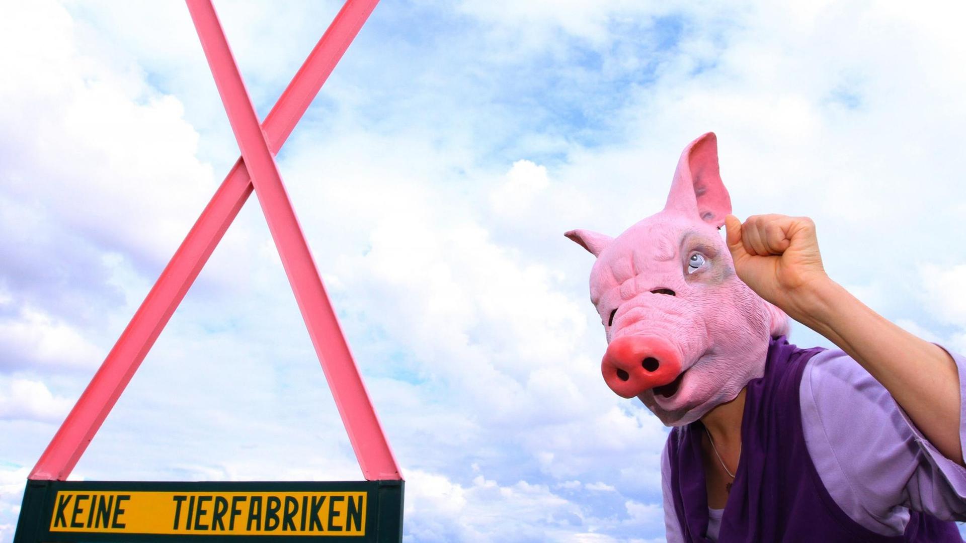 Eine Person trägt eine Schweinemaskte und ballt die Faust kämpferisch. Daneben ein pinke X welches die Aufschrift "Keine Tierfabriken" trägt. Eine Person trägt eine Schweinemaskte und ballt die Faust kämpferisch. Daneben ein pinke X welches die Aufschrift "Keine Tierfabriken" trägt.