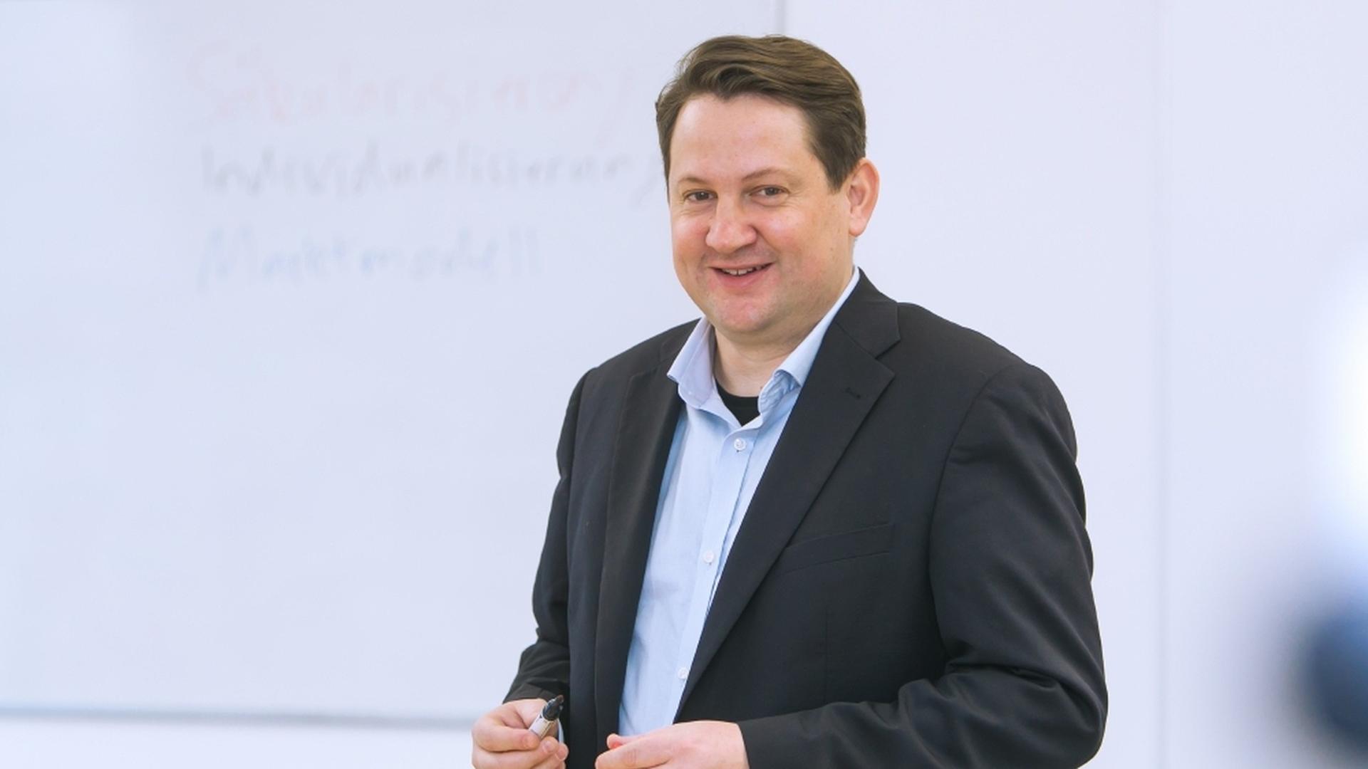 Dr. phil. Alexander Yendell, steht vor einer weißen Wand und lächelt in die Kamera, er hat einen Anzug an und einen Stift in der Hand. Dr. phil. Alexander Yendell, steht vor einer weißen Wand und lächelt in die Kamera, er hat einen Anzug an und einen Stift in der Hand.