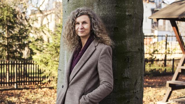 Eine Fotografie der Schriftstellerin Anna Kaleri. Sie steht in einem braunen Mantel mit brombeerfarbenen Schal vor dem dicken Stamm eines Baumes im Herbst.