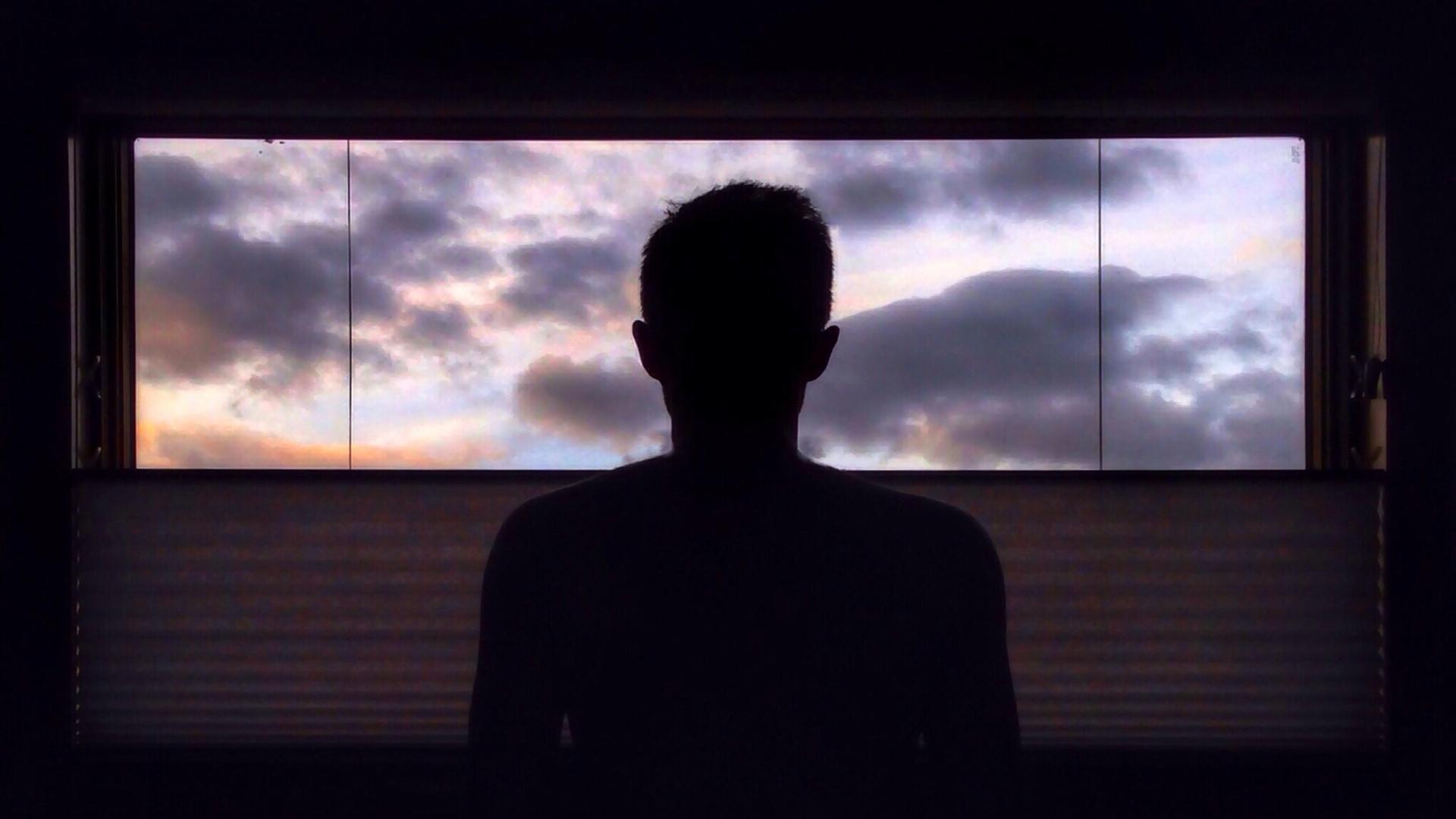 Eine Person sitzt in einem dunklen Raum vor einem Fenster, durch das ein dramatisch bewölkter Himmel zu sehen ist. Eine Person sitzt in einem dunklen Raum vor einem Fenster, durch das ein dramatisch bewölkter Himmel zu sehen ist.