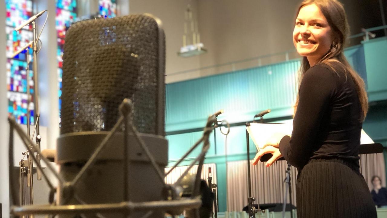 Eine junge Frau schaut über ihre Schulter, im Vordergrund ein großes Mikrophon. Eine junge Frau schaut über ihre Schulter, im Vordergrund ein großes Mikrophon.