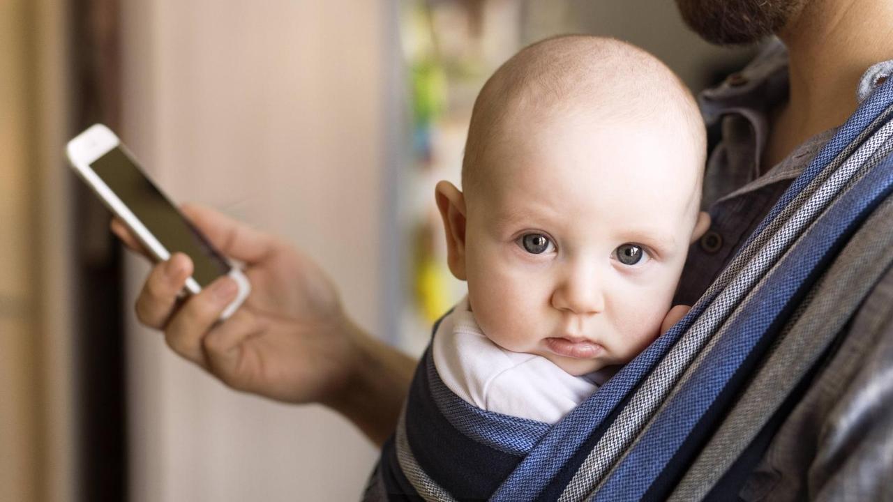 Ein Vater trägt ein Baby an der Brust und schaut dabei auf sein Smartphone Ein Vater trägt ein Baby an der Brust und schaut dabei auf sein Smartphone
