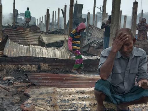 Vereinzelte Menschen laufen durch ein Gelände mit abgebrannten Hütten. Im Vordergrund ein älterer Mann, der auf einem Stück Wellblech sitzt und den Kopf in die Hand gestützt hat.