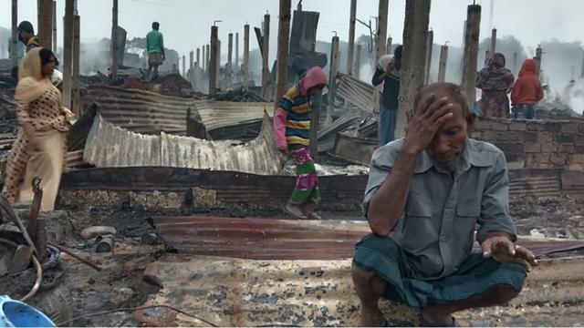 Vereinzelte Menschen laufen durch ein Gelände mit abgebrannten Hütten. Im Vordergrund ein älterer Mann, der auf einem Stück Wellblech sitzt und den Kopf in die Hand gestützt hat.
