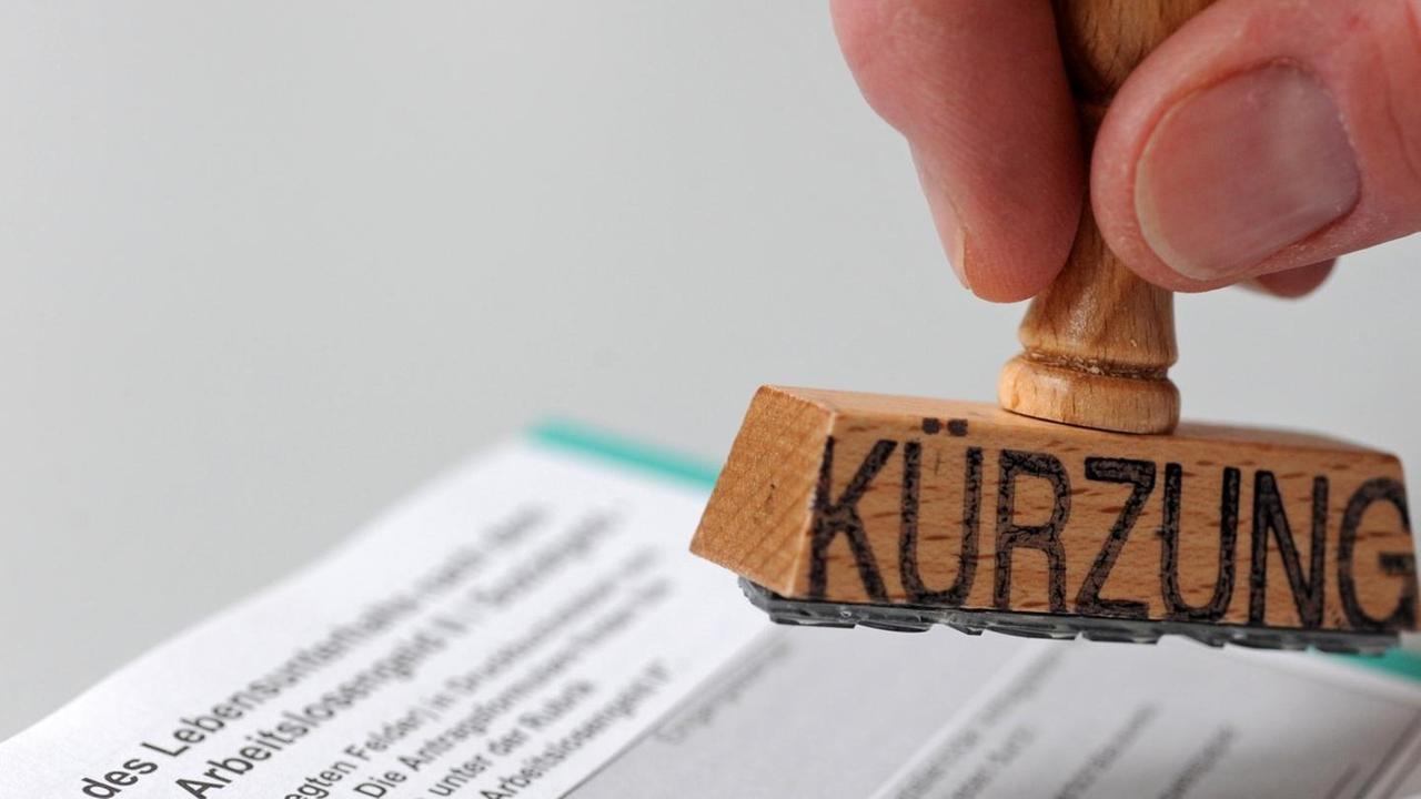 ILLUSTRATION - Ein Mann hält am Mittwoch (16.06.2010) einen Stempel, auf dem das Wort "Kürzung" steht, über ein Formular zur Beantragung von Arbeitslosengeld II (ALG II), umgangssprachlich auch "Hartz IV" genannt. Das Alg II ist die Grundsicherung für erwerbsfähige Hilfebedürftige nach dem Sozialgesetzbuch II (SGB II). Foto: Andreas Gebert dpa/lby | Verwendung weltweit ILLUSTRATION - Ein Mann hält am Mittwoch (16.06.2010) einen Stempel, auf dem das Wort "Kürzung" steht, über ein Formular zur Beantragung von Arbeitslosengeld II (ALG II), umgangssprachlich auch "Hartz IV" genannt. Das Alg II ist die Grundsicherung für erwerbsfähige Hilfebedürftige nach dem Sozialgesetzbuch II (SGB II). Foto: Andreas Gebert dpa/lby | Verwendung weltweit