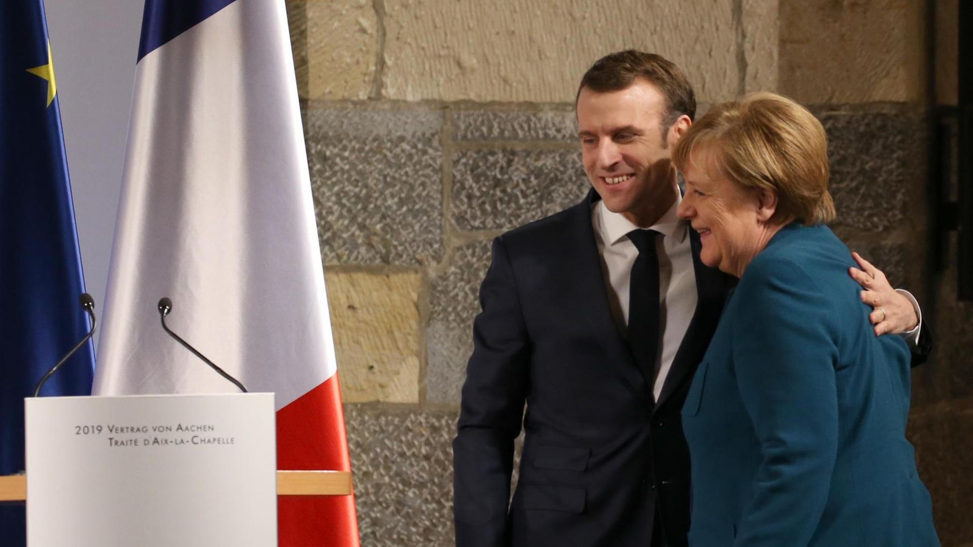 Präsident Macron umarmt Bundeskanzlerin Merkel. Neben ihnen stehen Flaggen.