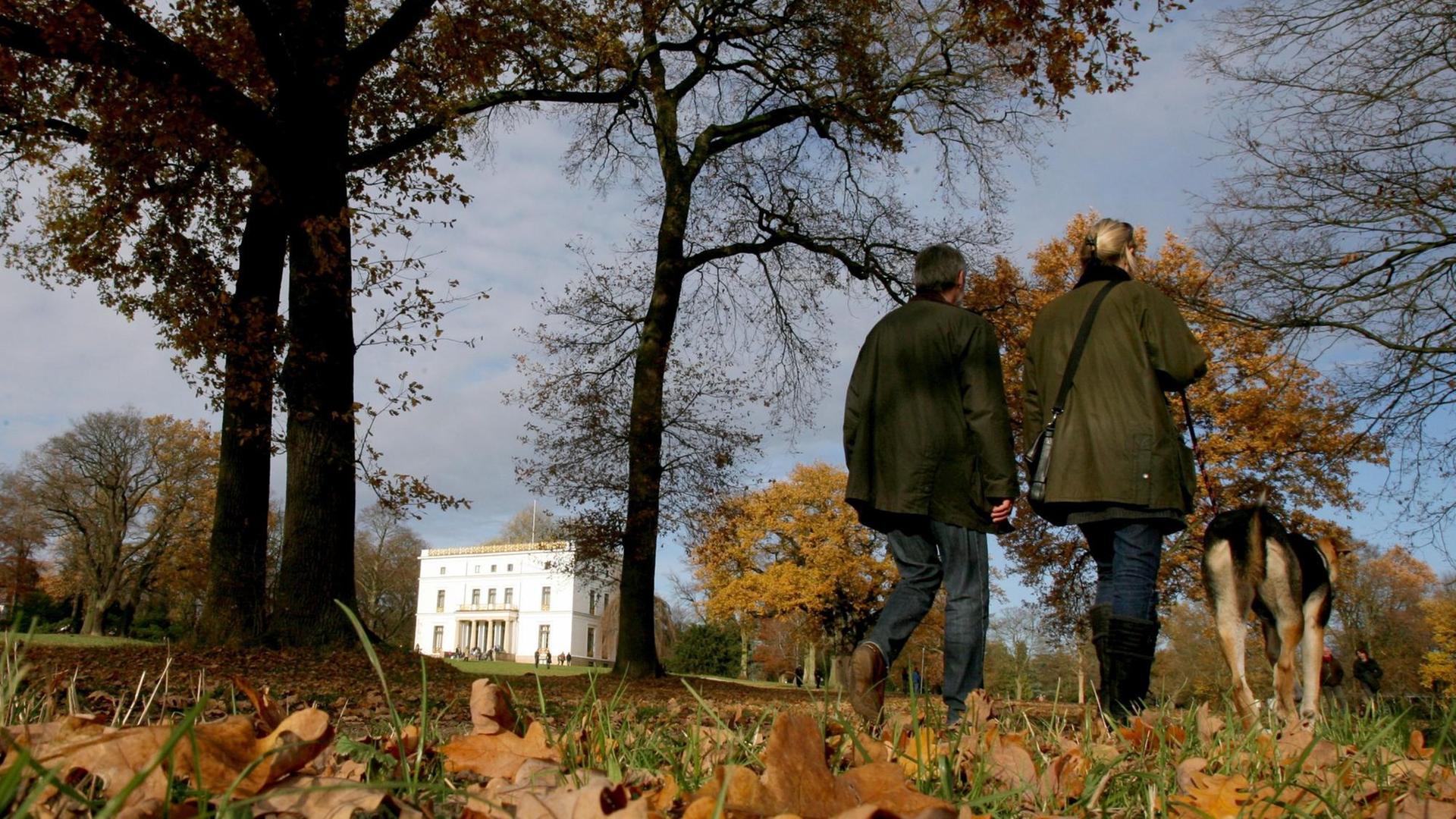 Zwei Spaziergänger gehen in Hamburg im Jenisch Park mit ihrem Hund spazieren, im Hintergrund steht das Jenisch Haus.