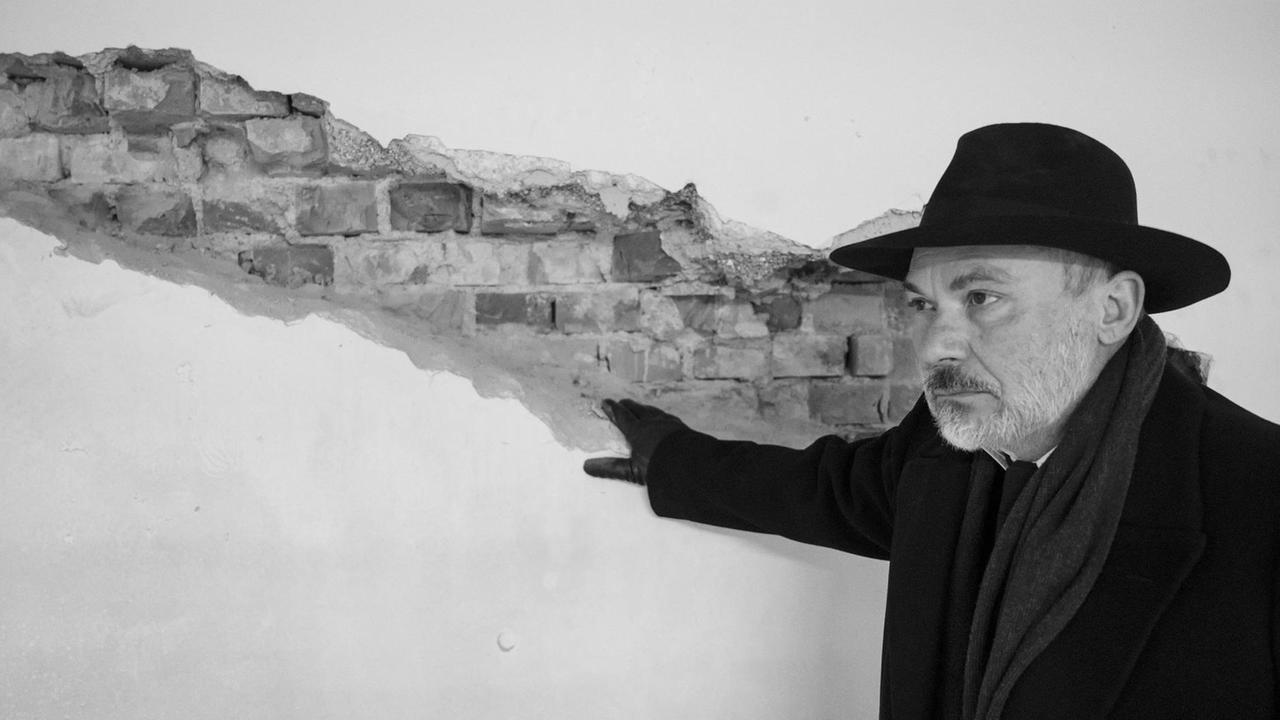 Der Schauspieler Klaus Müller steht mit Hut vor einer Wand, die zum Teil unverputzt ist. 