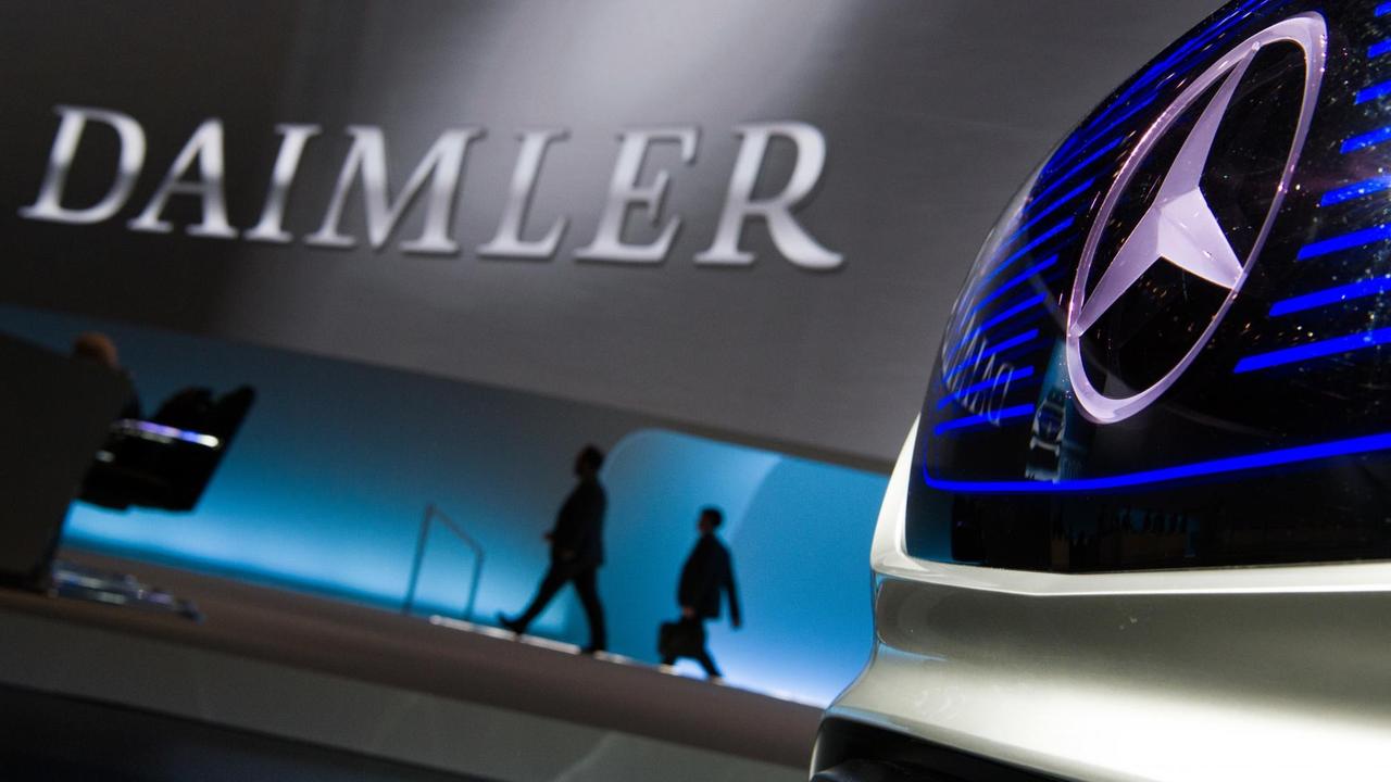 Im Vordergrund rechts ein Mercedes-Logo, im Hintergrund der Schriftzug Daimler an einer Wand, davor Silhouetten von Menschen.