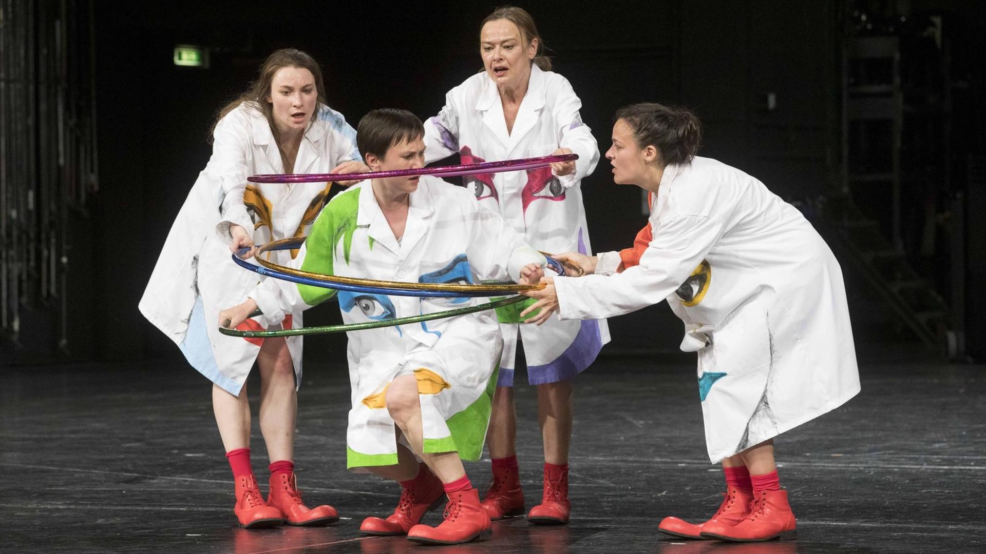 Eine Gruppe Schauspielerinnen in weißen Kostümen legt bunte Plastikringe über eine weitere Schauspielerin. Eine Gruppe Schauspielerinnen in weißen Kostümen legt bunte Plastikringe über eine weitere Schauspielerin.