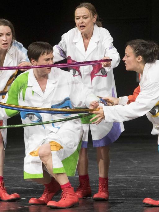 Eine Gruppe Schauspielerinnen in weißen Kostümen legt bunte Plastikringe über eine weitere Schauspielerin.