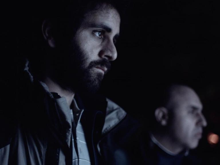 Filmsszene aus dem Kurzfil "Proll!. Murat (Erol Afsin) steht nehen einer weiteren Person im Dunkeln und schaut zur Seite in Richtung des Lichts.