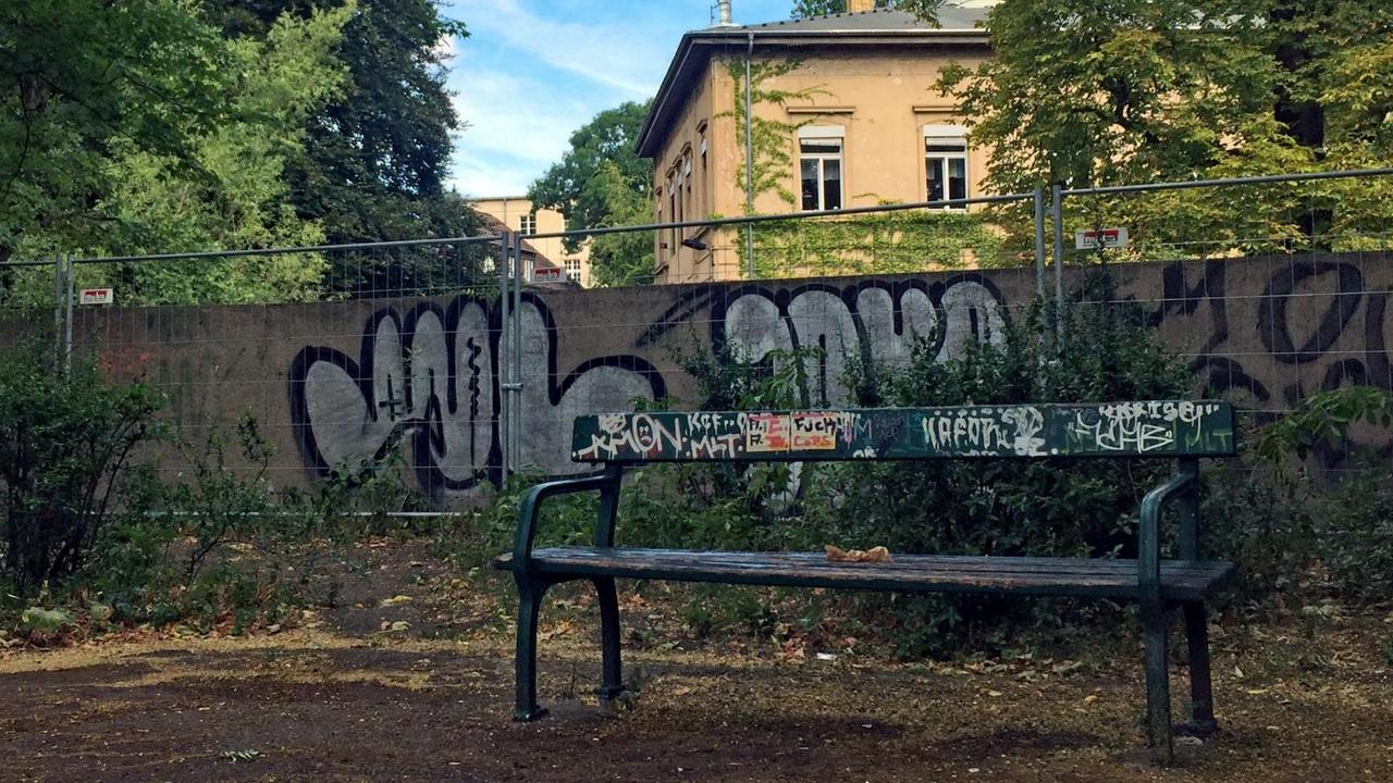 Eine heruntergekommene Parkbank steht vor einem Bauzaun, der eine mit Graffiti besprühte Mauer abschirmt, hinter der ein Gebäude hervorragt. Eine heruntergekommene Parkbank steht vor einem Bauzaun, der eine mit Graffiti besprühte Mauer abschirmt, hinter der ein Gebäude hervorragt.