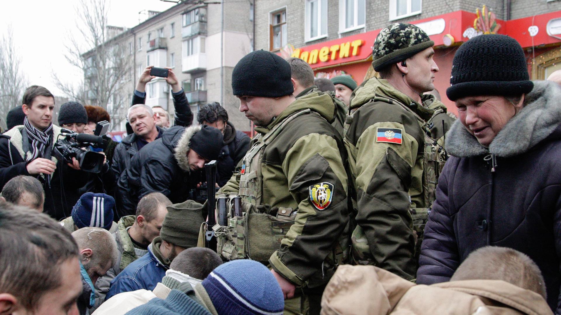 Menschenmenge in einer Stadt, darin knien einige Kriegsgefangene auf dem Boden, vor ihnen Männer in tarnfarbenen Uniformen. Menschenmenge in einer Stadt, darin knien einige Kriegsgefangene auf dem Boden, vor ihnen Männer in tarnfarbenen Uniformen.