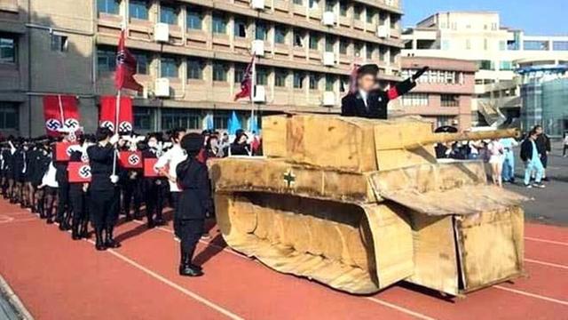 Ein Papp-Panzer auf einer Sportbahn, darauf zeigt ein Mann den Hitlergruß. Neben und hinter dem Panzer marschieren Schüler in Nazi-Uniform.