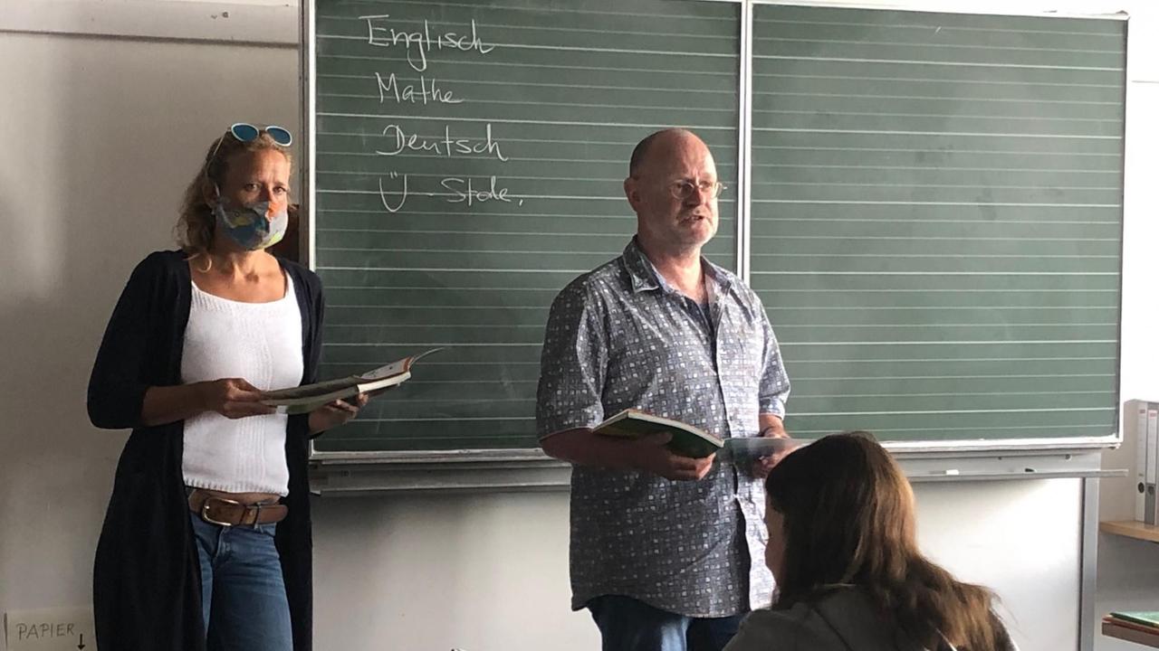 Vor der Tafel in einem Klassenzimmer stehen ein Lehrer und eine Lehrerin mit jeweils einem aufgeschlagenen Schulbuch in der Hand. Die Lehrerin trägt einen Mundschutz. Vor der Tafel in einem Klassenzimmer stehen ein Lehrer und eine Lehrerin mit jeweils einem aufgeschlagenen Schulbuch in der Hand. Die Lehrerin trägt einen Mundschutz.