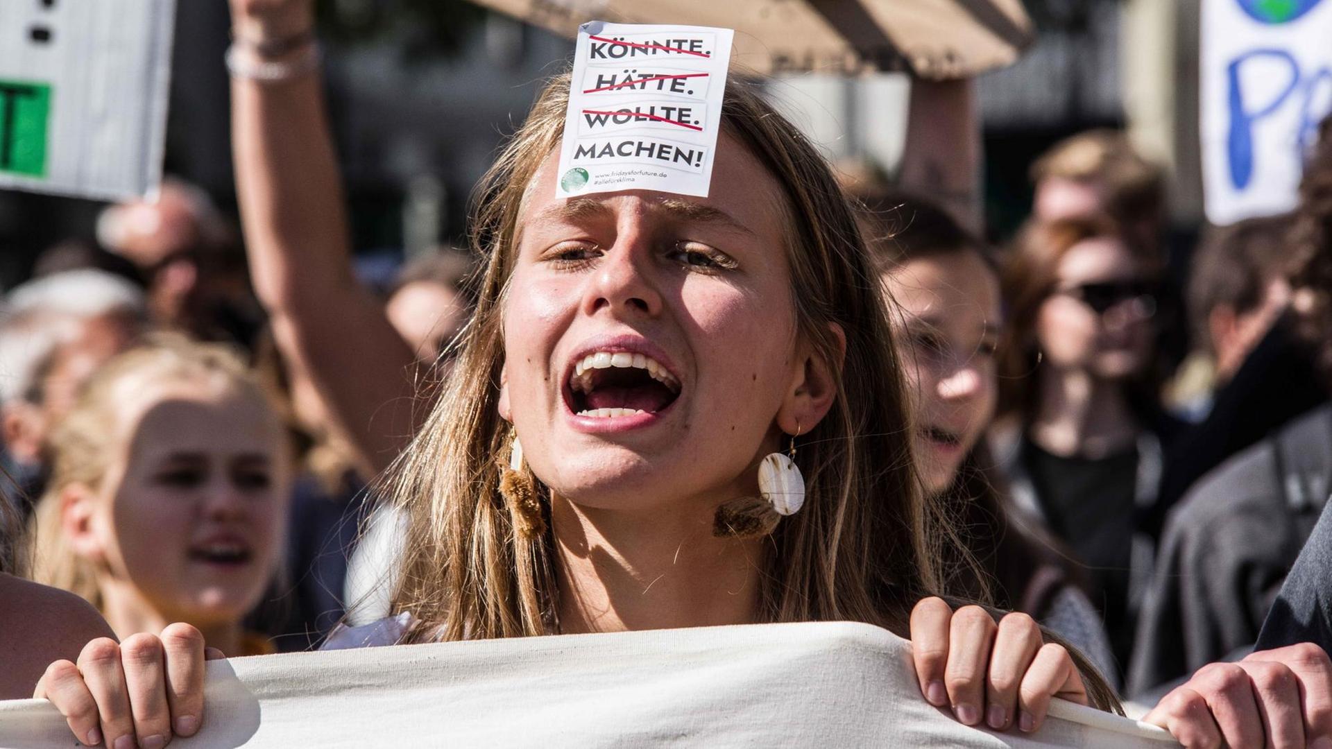 Eine junge Frau mit einem offenen Mund steht inmitten des Demonstrationszuges der fridays for future, einen Flyer auf ihrer Stirn klebend. Die Wörter könnte, hätte, wollte stehen durchgestrichen darauf und das Wort machen mit einem Ausrufezeichen dahinter. Eine junge Frau mit einem offenen Mund steht inmitten des Demonstrationszuges der fridays for future, einen Flyer auf ihrer Stirn klebend. Die Wörter könnte, hätte, wollte stehen durchgestrichen darauf und das Wort machen mit einem Ausrufezeichen dahinter.