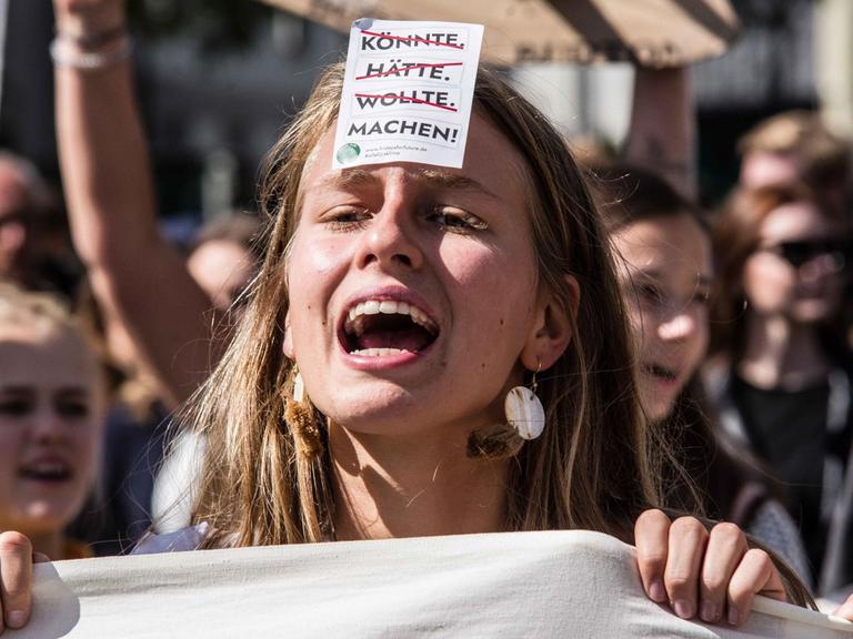 Eine junge Frau mit einem offenen Mund steht inmitten des Demonstrationszuges der fridays for future, einen Flyer auf ihrer Stirn klebend. Die Wörter könnte, hätte, wollte stehen durchgestrichen darauf und das Wort machen mit einem Ausrufezeichen dahinter.