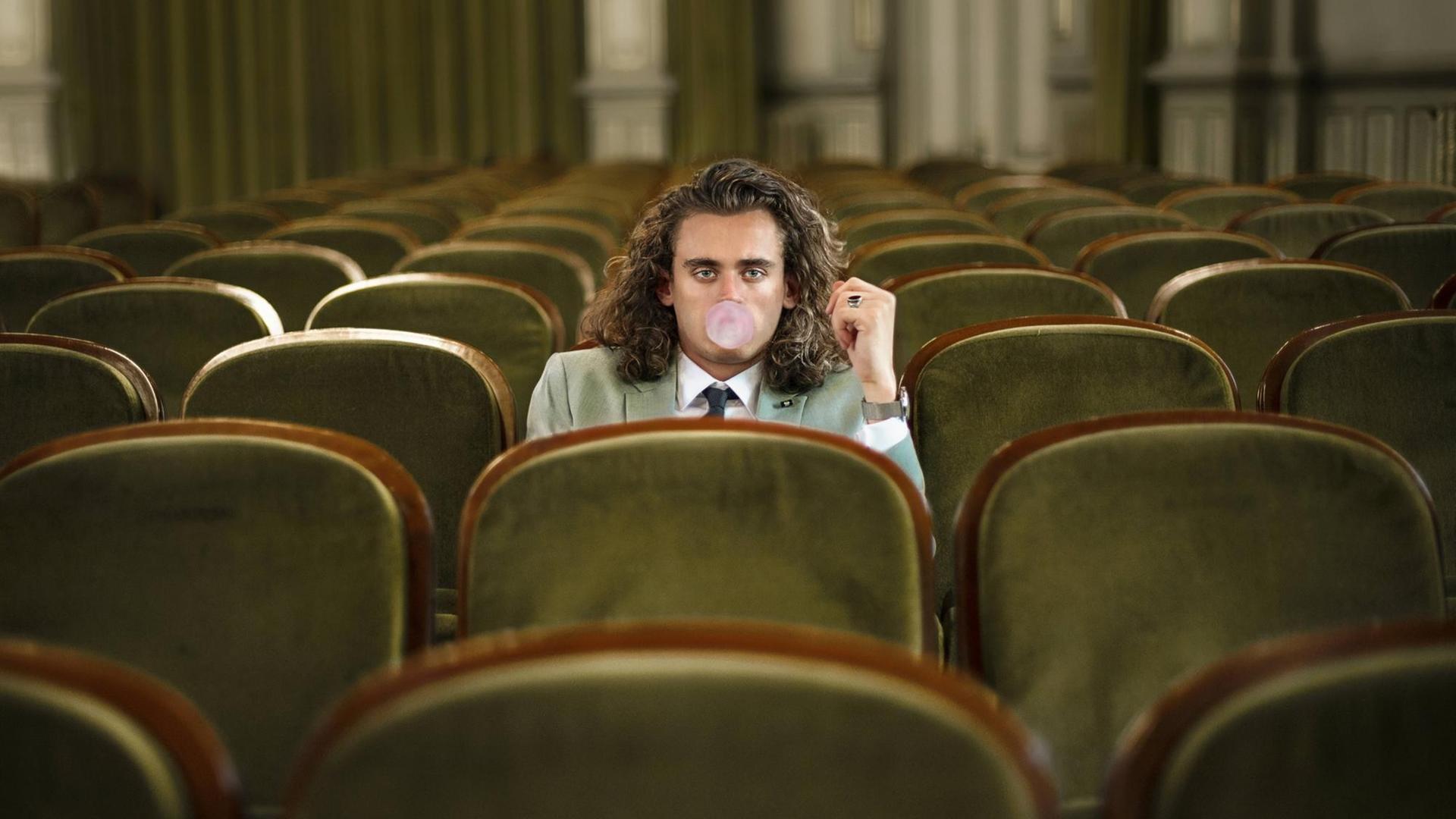 Ein gelangweilter junger Mann sitzt allein in einem Theatersaal und macht eine Kaugummiblase. Ein gelangweilter junger Mann sitzt allein in einem Theatersaal und macht eine Kaugummiblase.