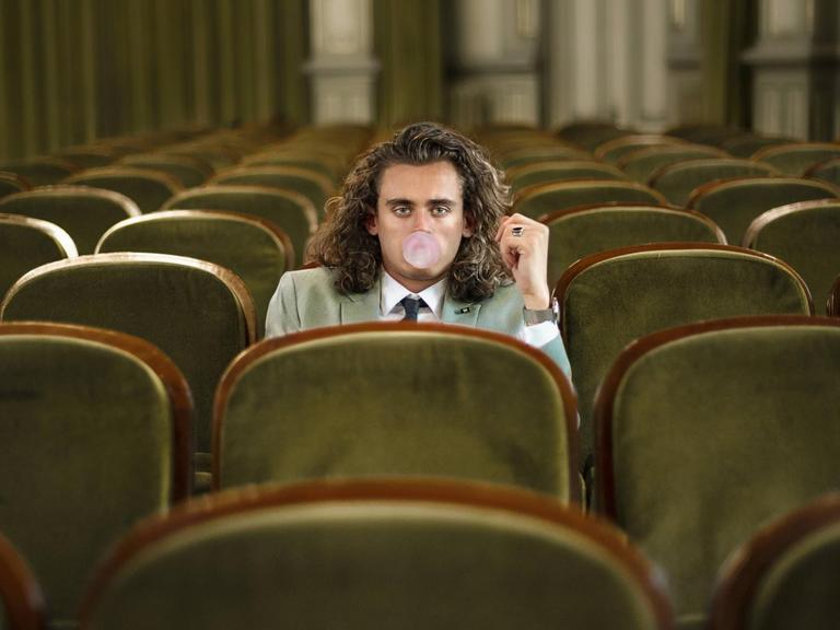 Ein gelangweilter junger Mann sitzt allein in einem Theatersaal und macht eine Kaugummiblase.