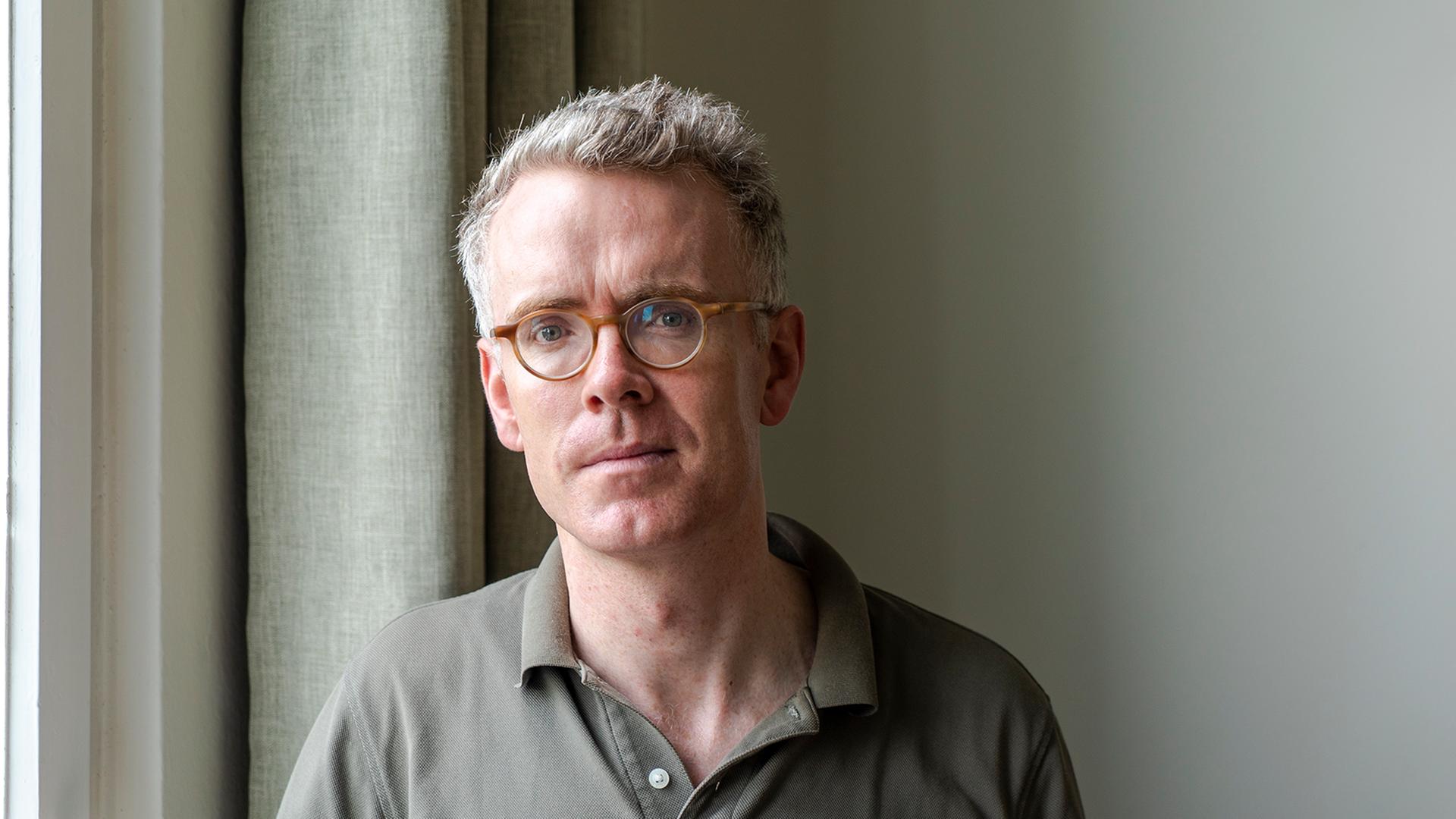 Derek Scally trägt Brille, steht am Fenster und schaut in die Kamera. Derek Scally trägt Brille, steht am Fenster und schaut in die Kamera.