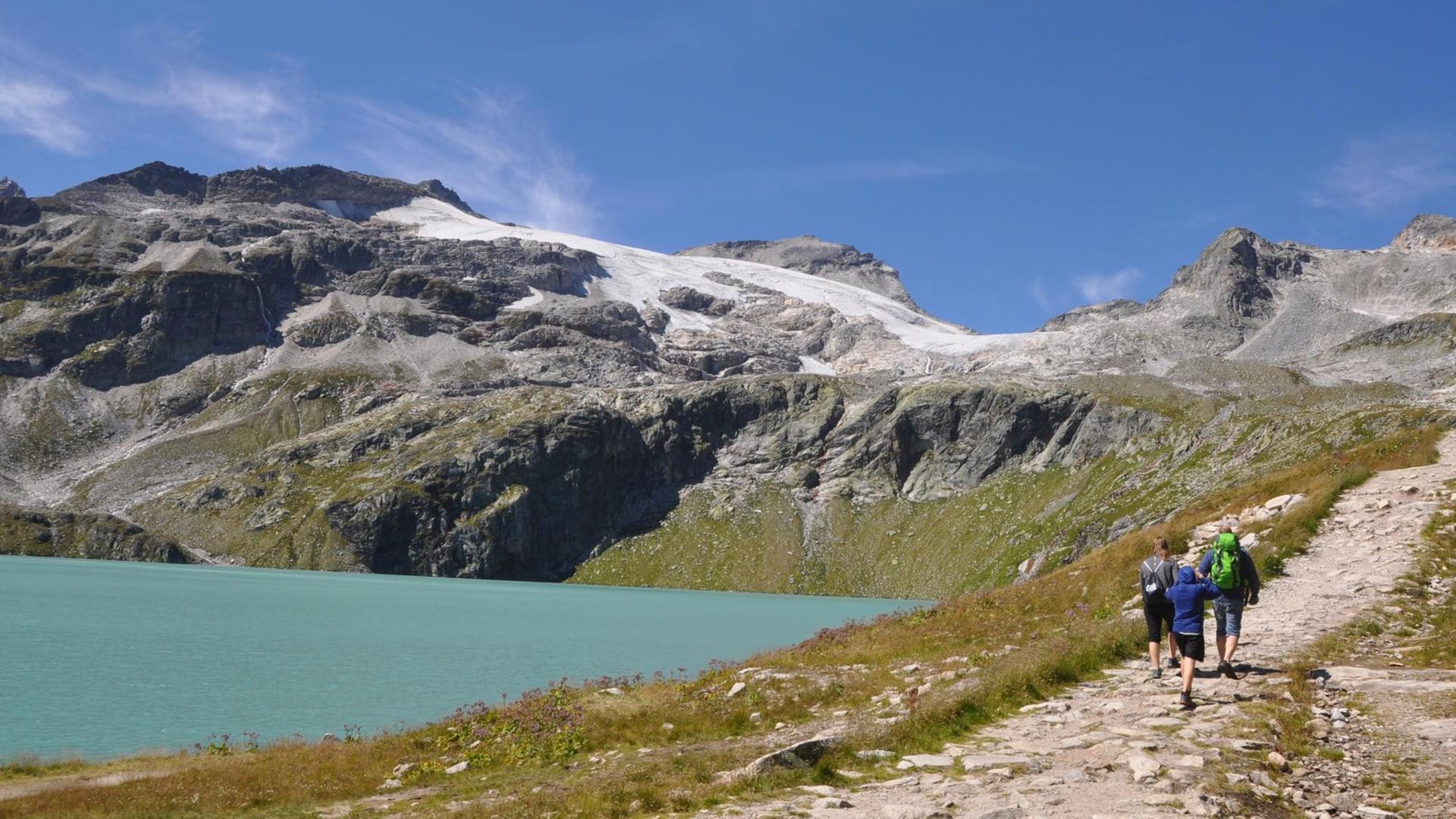 Abgebildet sind ein Bergsee mit einem Gletscher im Hintergrund, im Vordergrund gehen Wanderer Abgebildet sind ein Bergsee mit einem Gletscher im Hintergrund, im Vordergrund gehen Wanderer