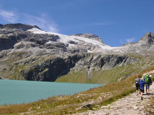 Abgebildet sind ein Bergsee mit einem Gletscher im Hintergrund, im Vordergrund gehen Wanderer
