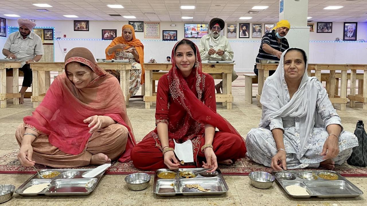 Drei Frauen in roten und weißen Saris und mit verhülltem Haar sitzen nebeneinander auf einem rot gemusterten Teppich in einer großen Halle, vor ihnen auf dem Boden stehen Tabletts mit Essen, hinter ihnen in einiger Entferung sitzen Männer und Frauen an Tischen und essen ebenfalls. Drei Frauen in roten und weißen Saris und mit verhülltem Haar sitzen nebeneinander auf einem rot gemusterten Teppich in einer großen Halle, vor ihnen auf dem Boden stehen Tabletts mit Essen, hinter ihnen in einiger Entferung sitzen Männer und Frauen an Tischen und essen ebenfalls.