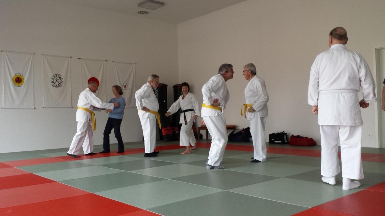 In einem Dojo stehen mehrere Gruppen. Die Menschen tragen zum Teil weiße Karateanzüge.